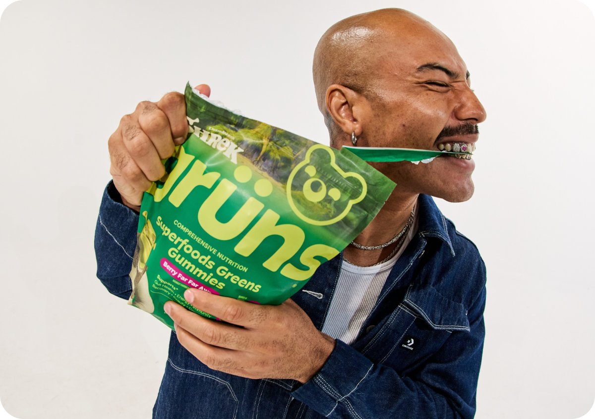 A bald man with a mustache and beard, wearing a denim jacket, uses his teeth to tear open a green "Grüns Superfoods Greens Gummies" pouch, eyes squeezed shut mid-bite.