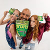 Three people posing playfully together, each holding up a green Shrek Grüns Superfoods Greens Gummies package, with the large main pack labeled Berry Far Far Away held in the center.