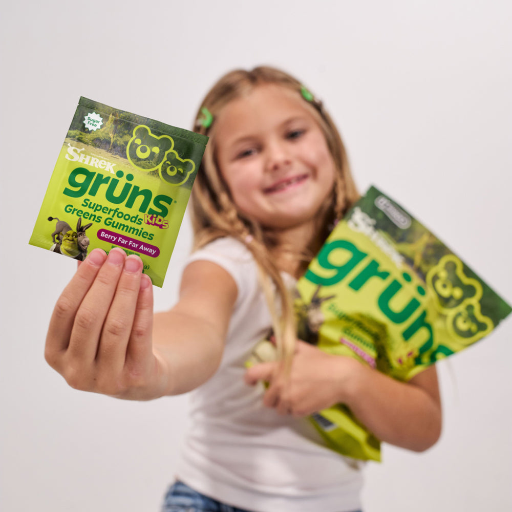Young girl smiling and holding up a Shrek Kids Grüns Superfoods Greens Gummies packet labeled Berry Far Far Away, with another green Grüns package in her other hand, slightly out of focus in the background.