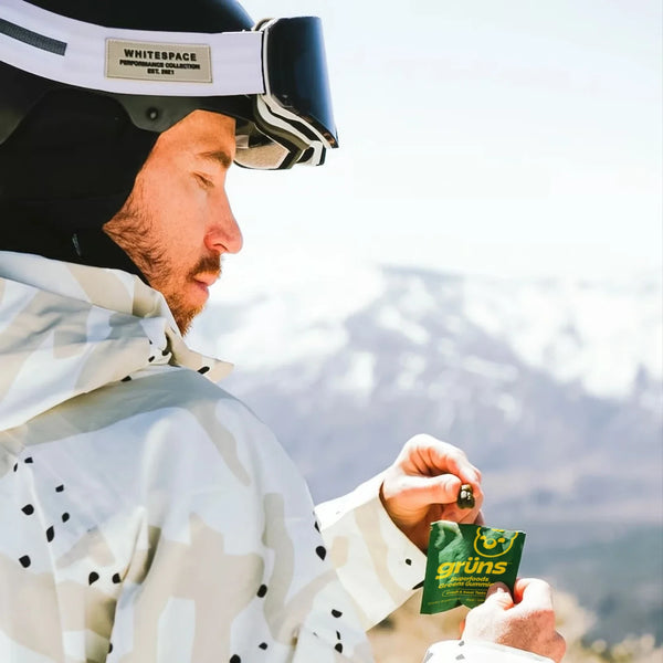 Shaun White in ski gear holding a green 'Grüns' snack packet with mountains in the background