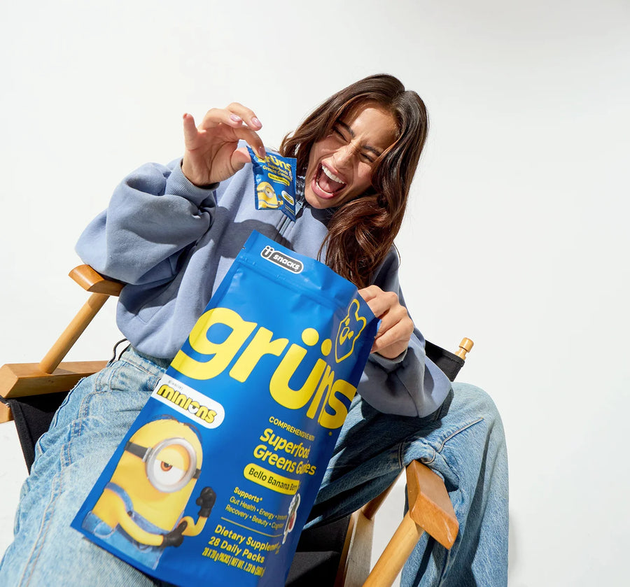 A woman sitting in a director's chair laughing while holding a Grüns Superfoods Greens Gummies bag with Minions branding and a single-serving pouch in her other hand.