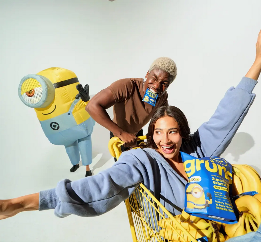 Two people playfully riding and pushing a shopping cart filled with bananas and a blue "Grüns Superfoods Greens Gummies" bag, with a person in an inflatable Minion costume standing beside them against a plain background.