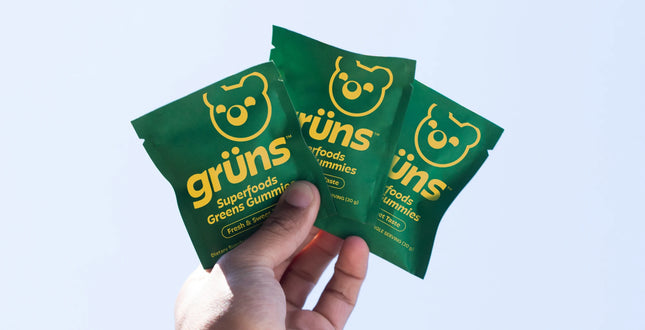 Three green packets of grüsns superfoods greens gummies held by a hand against a light blue background.