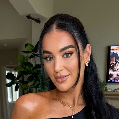 A woman with long dark hair pulled back poses for a close-up portrait indoors, wearing a black top, earrings, and layered necklaces, with a plant and television visible in the background.
