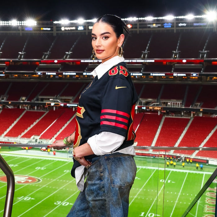 A woman poses inside a football stadium wearing a dark football jersey over a white shirt and jeans, with the brightly lit field and empty red stadium seats visible behind her.
