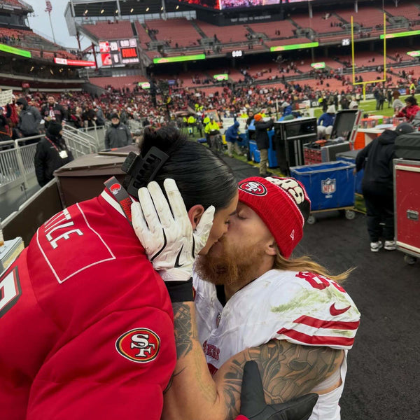 A couple kisses on the field at a football stadium, with the woman wearing a red jersey and the man wearing a white uniform and red team beanie. Fans and stadium seating are visible in the background.
