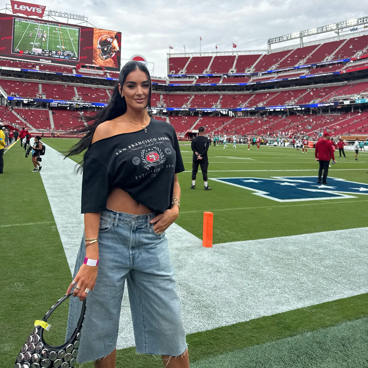 A woman poses on the field at Levi’s Stadium wearing a black San Francisco 49ers cropped T-shirt and denim shorts, holding a black handbag, with the football field, red stadium seats, and scoreboard visible in the background.