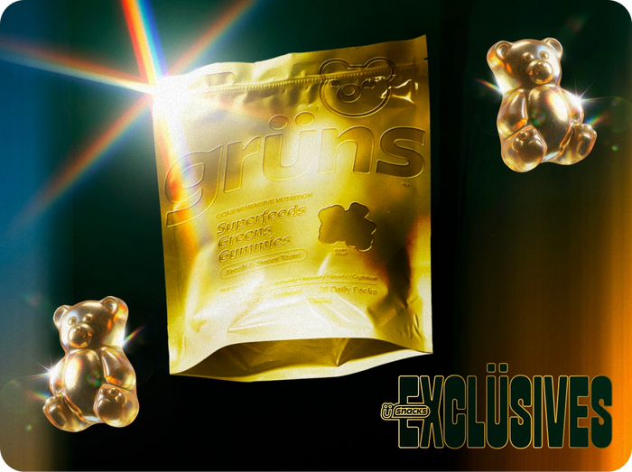 Dramatic product shot of a metallic gold grüns Superfoods Greens Gummies pouch bag lit with a rainbow lens flare, flanked by two shiny gold gummy bear figures, with a "snacks Exclüsives" logo in the bottom right corner.