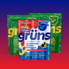 Three Grüns Superfoods Greens Gummies pouches on a blue-to-red gradient background, with a red, white, and blue Popsicle Firecracker pouch in front of two green pouches with yellow branding, featuring a blue bear-face logo in a top hat, an illustration of red, white, and blue popsicles, and star-shaped gummies with fruit accents.