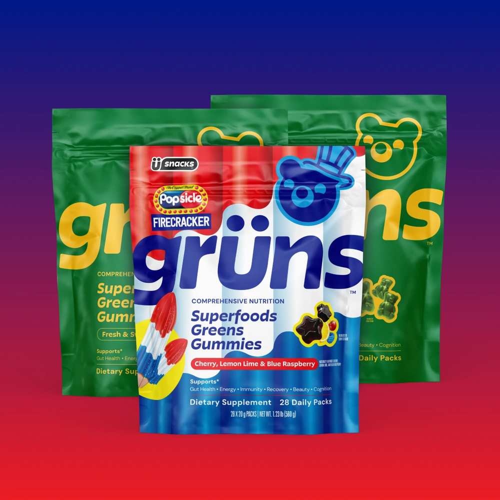Three Grüns Superfoods Greens Gummies pouches on a blue-to-red gradient background, with a red, white, and blue Popsicle Firecracker pouch in front of two green pouches with yellow branding, featuring a blue bear-face logo in a top hat, an illustration of red, white, and blue popsicles, and star-shaped gummies with fruit accents.