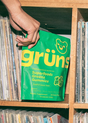 A hand holding a green bag of Superfoods Greens Gummies in a shelf with records.