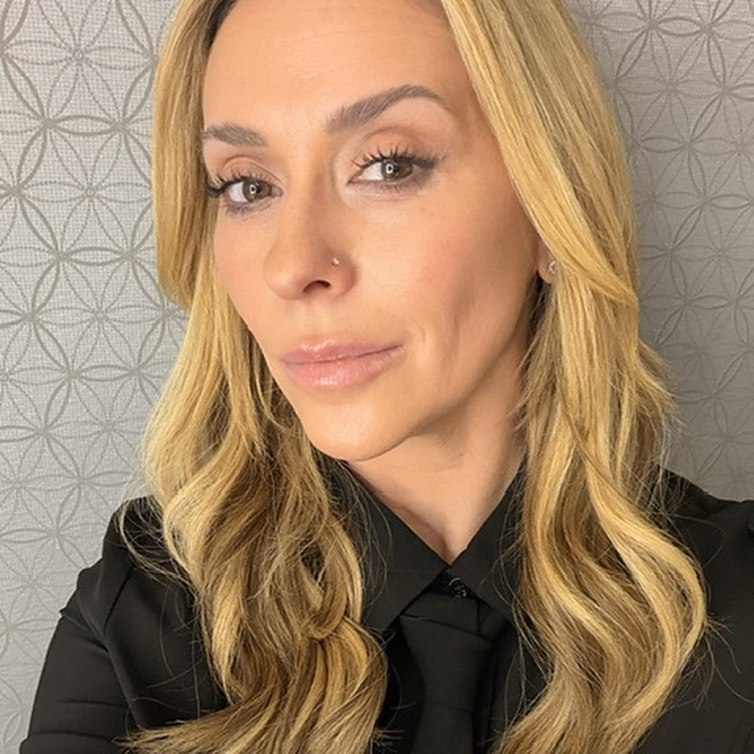 A close-up selfie of a woman with long wavy blonde hair, wearing a black button-up top with a tie-like neckline, against a gray patterned wallpaper background.