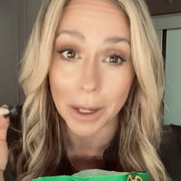 A close-up video-style shot of a woman with long wavy blonde hair talking to the camera, holding a gummy in one hand with a green Grüns bag visible at the bottom of the frame.