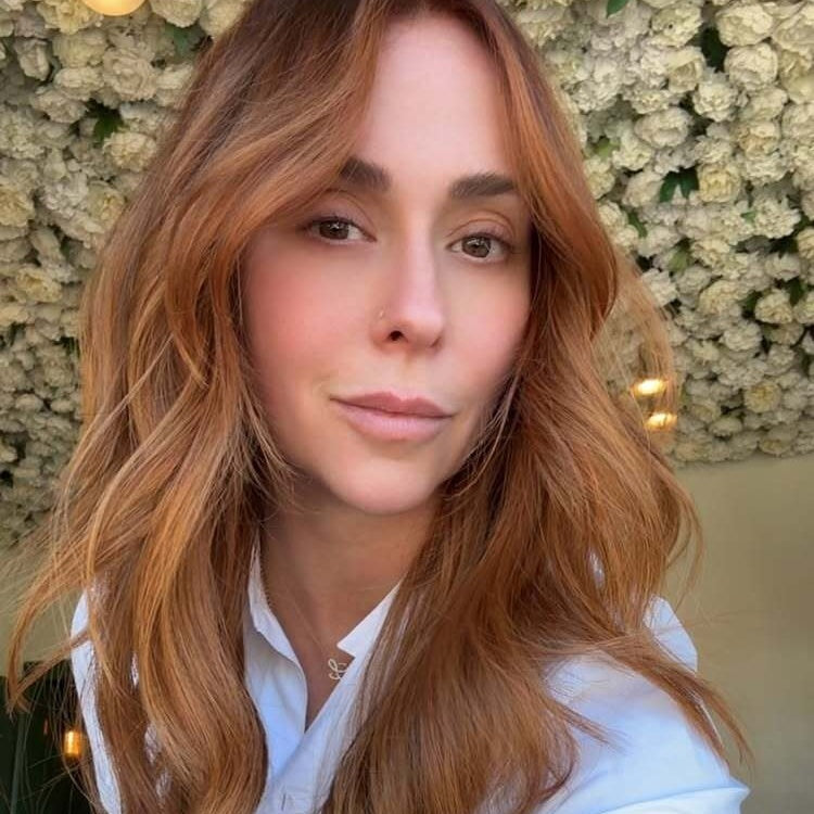 A close-up selfie of a woman with long wavy auburn hair and a nose piercing, wearing a white collared top with a small pendant necklace, standing in front of a wall covered in white flowers with warm string lights.