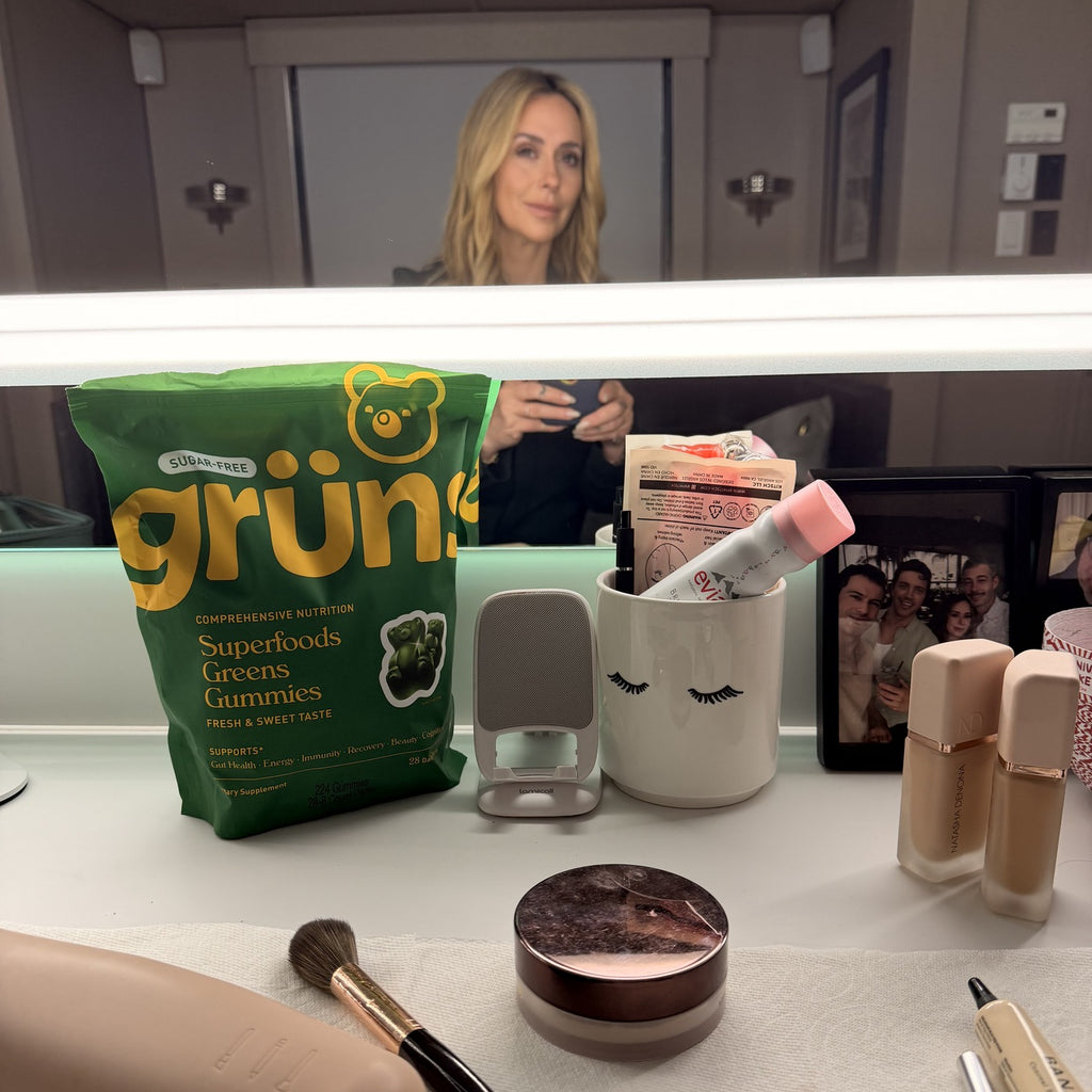 A green "Grüns Superfoods Greens Gummies" bag sitting on a vanity table surrounded by makeup items, with a woman with blonde wavy hair visible taking a photo in a lit mirror behind it.
