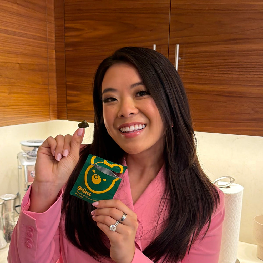 Woman smiling while holding up a green gummy and a green Grüns Superfoods Greens Gummies packet, in a kitchen with wood cabinets.