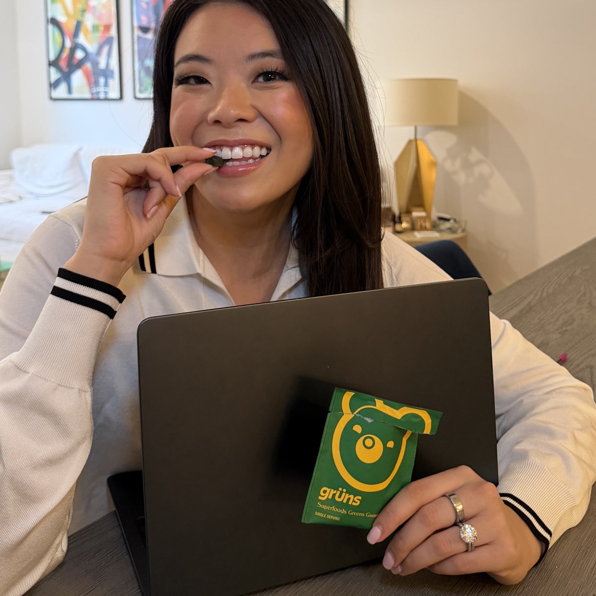 Woman smiling and eating a green gummy while sitting at a laptop, holding an open green Grüns Superfoods Greens Gummies packet.