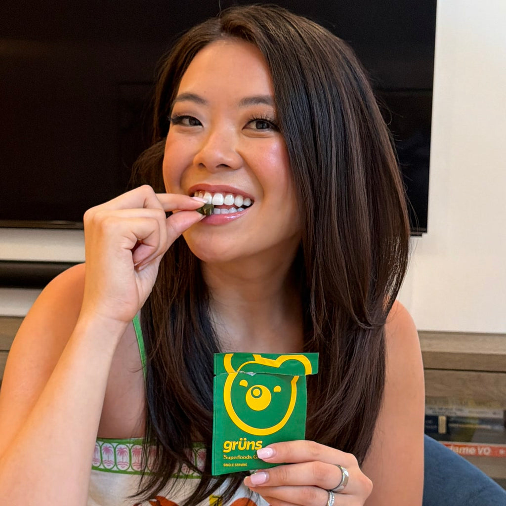 Woman smiling and eating a green gummy, holding an open green Grüns Superfoods Greens Gummies packet.