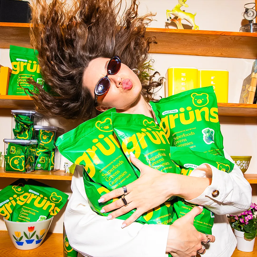 Person with sunglasses holding multiple green bags of gummies, hair flowing upwards.