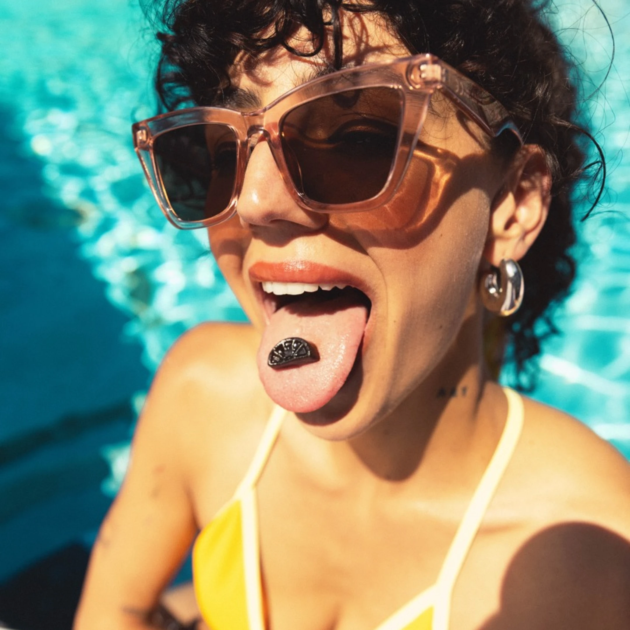 Person with sunglasses by a pool, holding a raspberry lemonade gummy on their tongue.