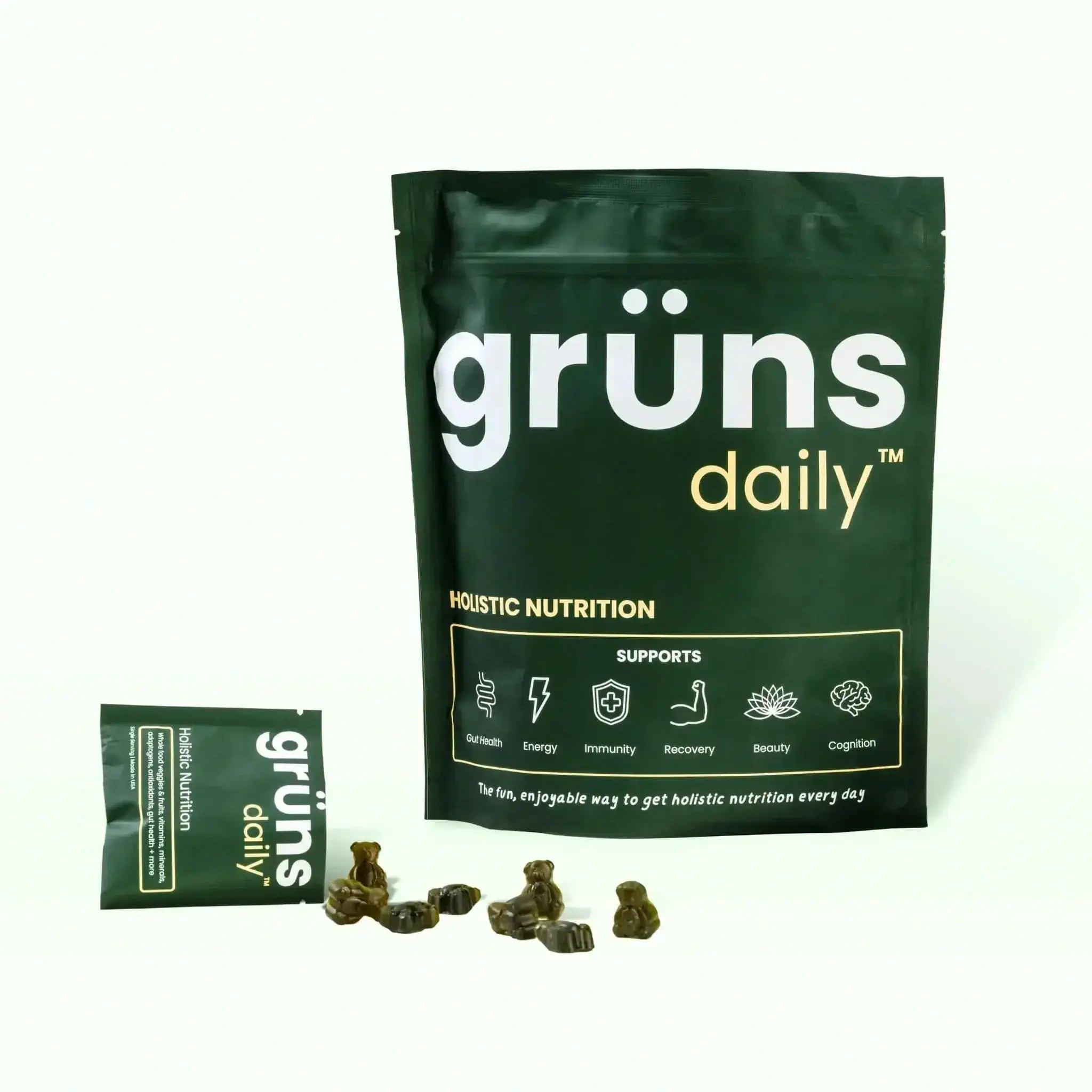 Packaging of 'grüns daily' holistic nutrition supplement with spilled supplements in the foreground.