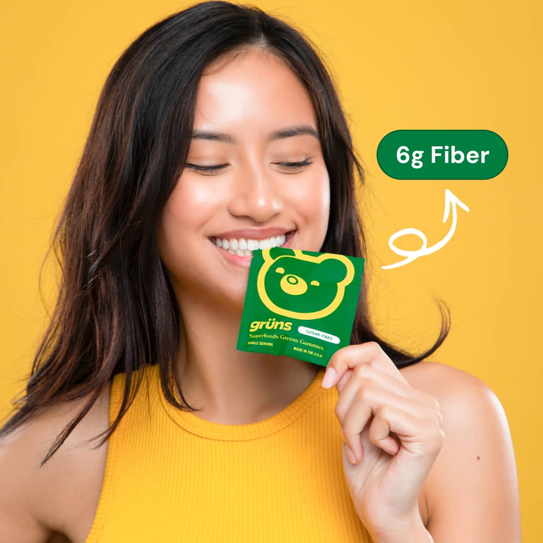 Smiling woman holding Grüns Superfoods Greens Gummies packet against yellow background.